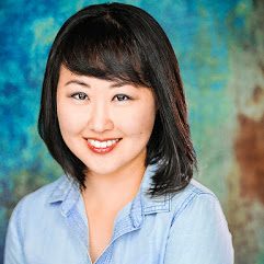 headshot of a woman with fair skin, shoulder-length black hair with swept bangs, wearing a chambray collared shirt, smiling at the camera in front of a teal and brown textured background. 