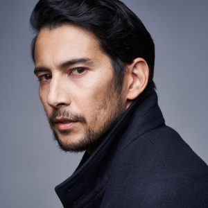 headshot of a man with brown skin and short black hair, brown eyes, and stubble on his face, looking over his left shoulder at the camera, wearing a black high-collared jacket, with grey background.